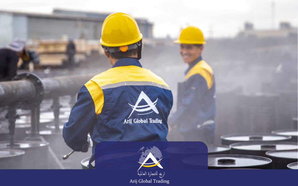 Two Arij Global Trading operational staff wearing branded blue and yellow uniforms and safety helmets overseeing the hot-filling process of export drums, with industrial steam visible on the active packaging line.