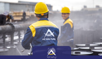 Two Arij Global Trading operational staff wearing branded blue and yellow uniforms and safety helmets overseeing the hot-filling process of export drums, with industrial steam visible on the active packaging line.