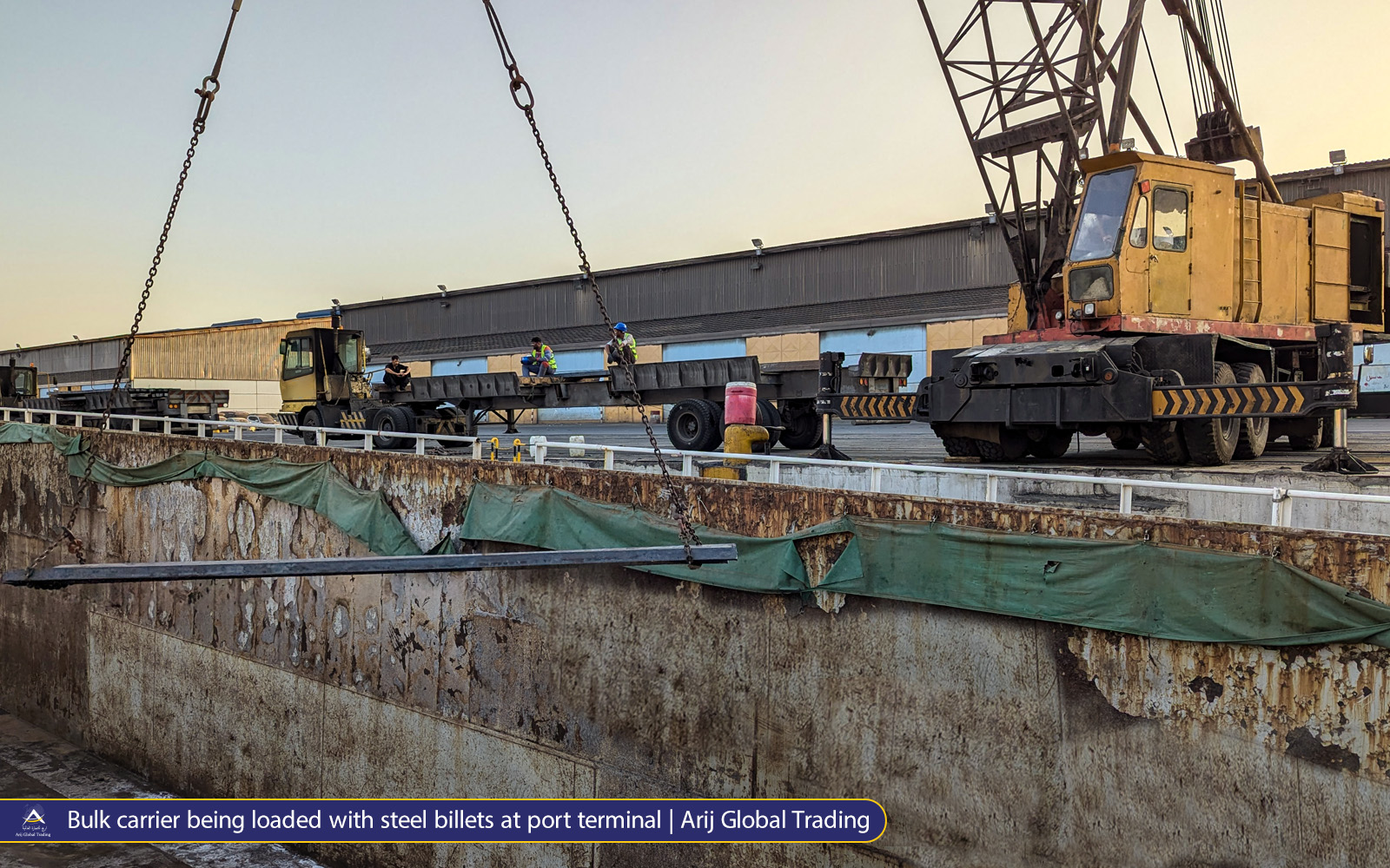 Bulk carrier loaded with Arijco Primmum steel billets at port terminal by orange crane and trucks, hi-vis workers managing operations