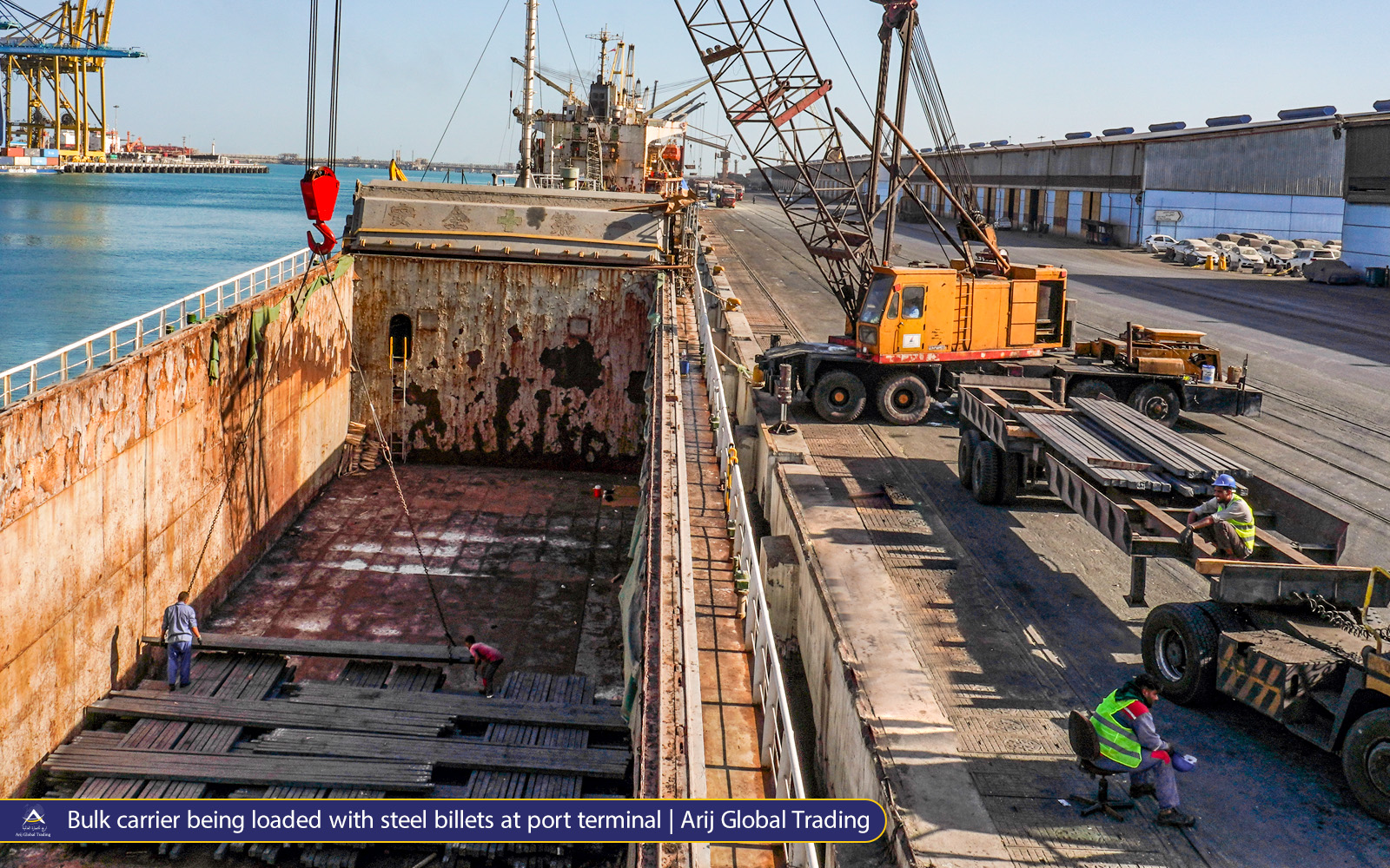 Bulk carrier being loaded with steel billets at port terminal Overhead view of workers loading stacks of Arijco Premium steel billets into the open hold of a bulk carrier using a yellow mobile crane, with trailers of steel on the quay beside a long warehouse at a busy seaport.