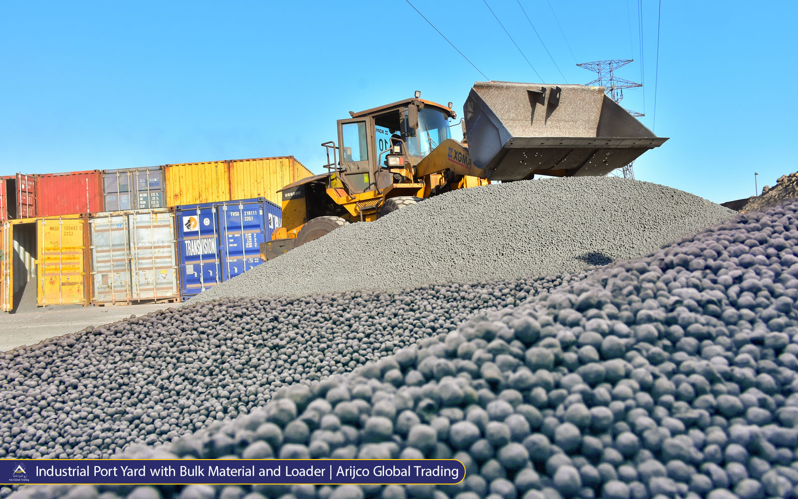 Industrial port yard with a yellow XGMA wheel loader behind piles of dark pellets, stacked shipping containers, and an Ark Global Trading banner.