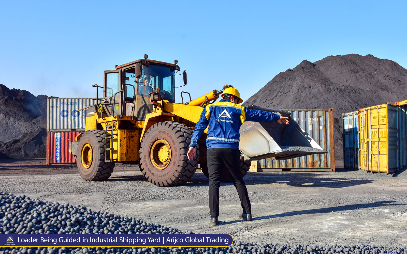 Worker pointing toward a yellow loader filled with dark pellets in a busy industrial yard with shipping containers and Arij Global Trading branding.