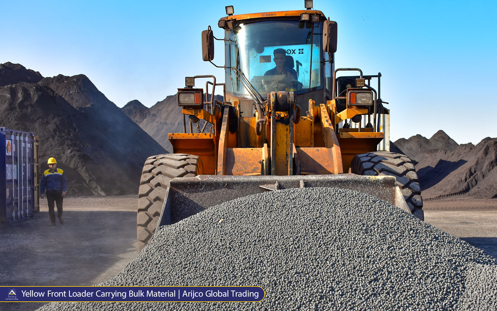 Yellow XGMA front loader filled with grey pellets in an industrial yard, with a worker beside a container and Arki Global Trading branding.